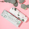 Rolled up Eucalyptus Print MakeUp Eraser next to packaging surrounded by eucalyptus leaves.