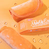 Two rolled up Juicy Orange MakeUp Eraser cloths next to packaging surrounded by waterdrops. 